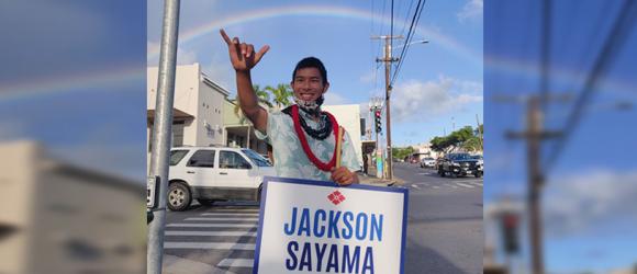 Sayama waves a sign under a rainbow